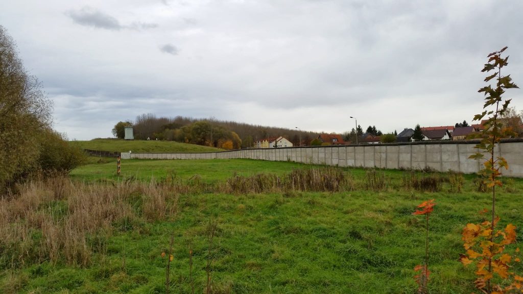 Ein Abschnitt der Mauer steht hinter einer Wiese, hinter ihr ragen die Dächer einiger Häuser.