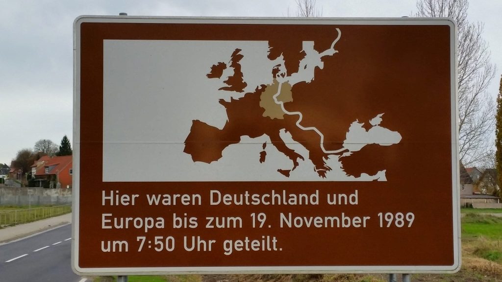 Braun weißes Schild, das eine Karte Europas zeigt, auf der die Mauer und Teilung eingezeichnet ist. darunter steht "Hier waren Deutschland und Europa bis zum 19. November 1989 um 7:50 Uhr geteilt."